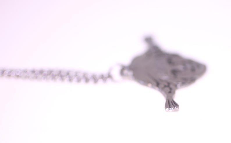 May include: A silver-toned pendant necklace featuring a detailed, textured manta ray design. The pendant is suspended from a delicate chain, creating a minimalist and elegant aesthetic. The necklace is set against a bright white background.