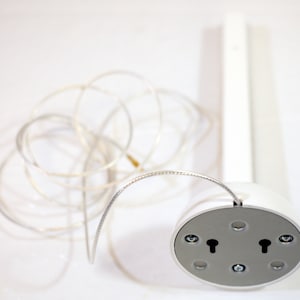 Puede incluir: Una lámpara de techo blanca con un cable plateado. La lámpara tiene una base redonda con dos ranuras en forma de ojo de cerradura y está unida a un soporte rectangular blanco.