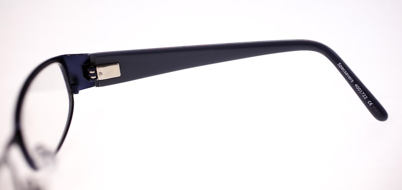 May include: A close-up of black eyeglasses. The frames have a silver rectangular detail on the temple. The arm of the glasses has the text "Specsavers 4001722 CE". The lenses are clear.