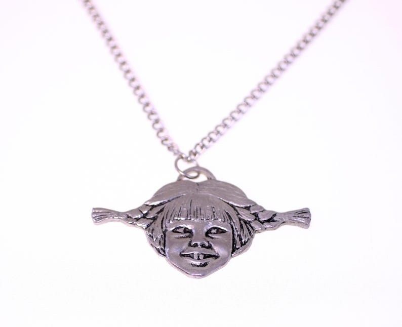 May include: A silver-toned pendant necklace featuring a detailed face with braided pigtails. The pendant is a sculpted design of a smiling girl's face. The necklace chain is a simple, linked design.