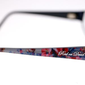 May include: Navy blue eyeglasses with a floral patterned arm in red, pink, and blue. The text "Red or Dead" is printed on the arm. The glasses have a classic rectangular shape. The frame is navy blue.