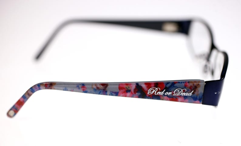 May include: Navy blue eyeglasses with a floral patterned arm in red, pink, and blue. The text "Red or Dead" is printed on the arm. The glasses have a classic rectangular shape. The frame is navy blue.
