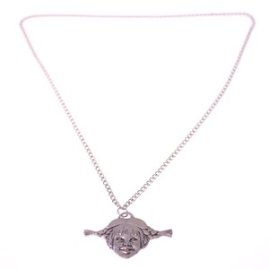 May include: A silver-toned necklace featuring a pendant shaped like a girl's face with braided pigtails. The pendant is suspended from a delicate chain. The background is plain white.