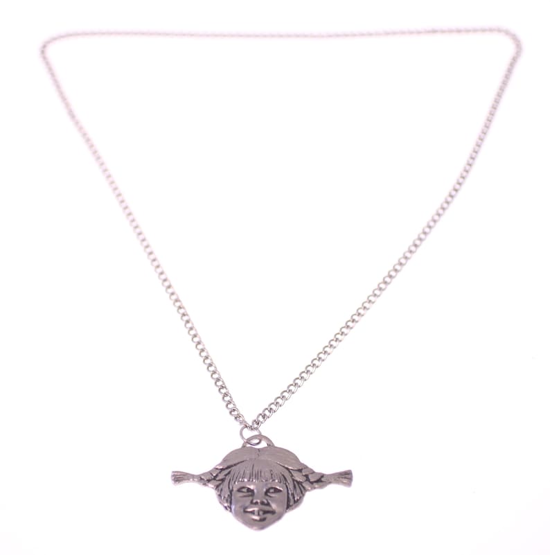 May include: A silver-toned necklace featuring a pendant shaped like a girl's face with braided pigtails. The pendant is suspended from a delicate chain. The background is plain white.