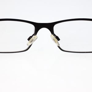 May include: Black rectangular eyeglasses with clear lenses. The frames are a matte black, and the temples are a translucent white. The nose pads are a light beige colour. The glasses are set against a white background.