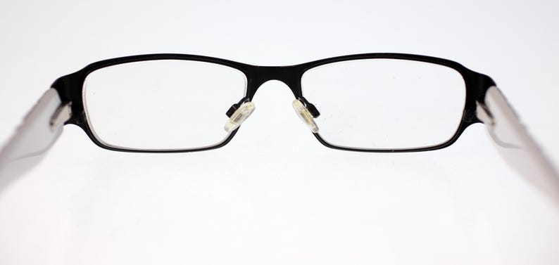 May include: Black rectangular eyeglasses with clear lenses. The frames are a matte black, and the temples are a translucent white. The nose pads are a light beige colour. The glasses are set against a white background.