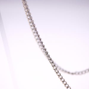 May include: Close-up of a silver-colored chain with a twisted rope design. The chain is angled diagonally across the frame against a white background. The links are small and closely connected.