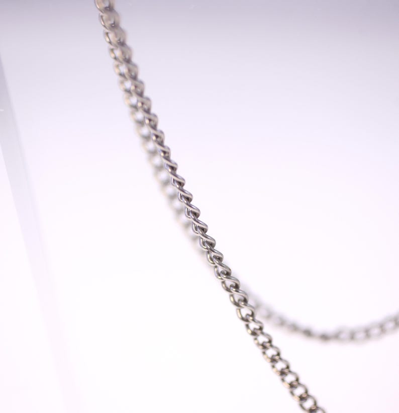 May include: Close-up of a silver-colored chain with a twisted rope design. The chain is angled diagonally across the frame against a white background. The links are small and closely connected.