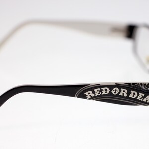 May include: Black-framed eyeglasses with the text "RED OR DEAD" on the temple. The arms and temples have white accents. The lenses are clear.