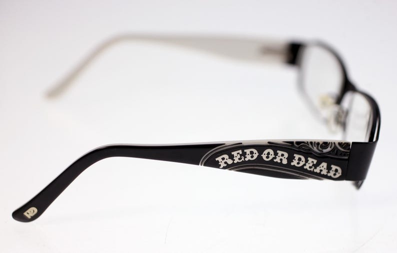 May include: Black-framed eyeglasses with the text "RED OR DEAD" on the temple. The arms and temples have white accents. The lenses are clear.