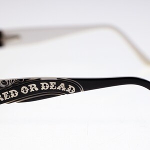 May include: Black framed eyeglasses with white arms. The words "RED OR DEAD" are printed in white on the black arm of the glasses. The glasses have a decorative design.