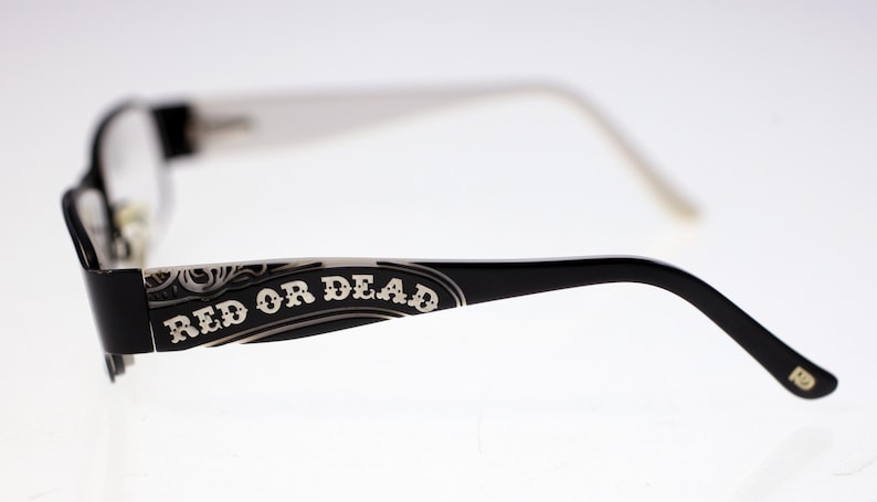 May include: Black framed eyeglasses with white arms. The words "RED OR DEAD" are printed in white on the black arm of the glasses. The glasses have a decorative design.