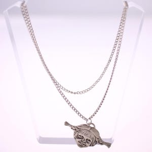 May include: A silver-toned necklace with a double chain design. The pendant features a detailed, stylized face with flowing hair. The necklace is displayed on a clear stand, showcasing its intricate details and design.