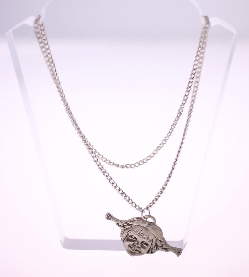 May include: A silver-toned necklace with a double chain design. The pendant features a detailed, stylized face with flowing hair. The necklace is displayed on a clear stand, showcasing its intricate details and design.