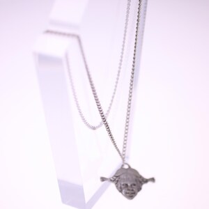 May include: A silver-colored necklace with a pendant shaped like a face with braided hair. The pendant is suspended from a delicate chain. The necklace is displayed on a clear, angled stand against a white background.