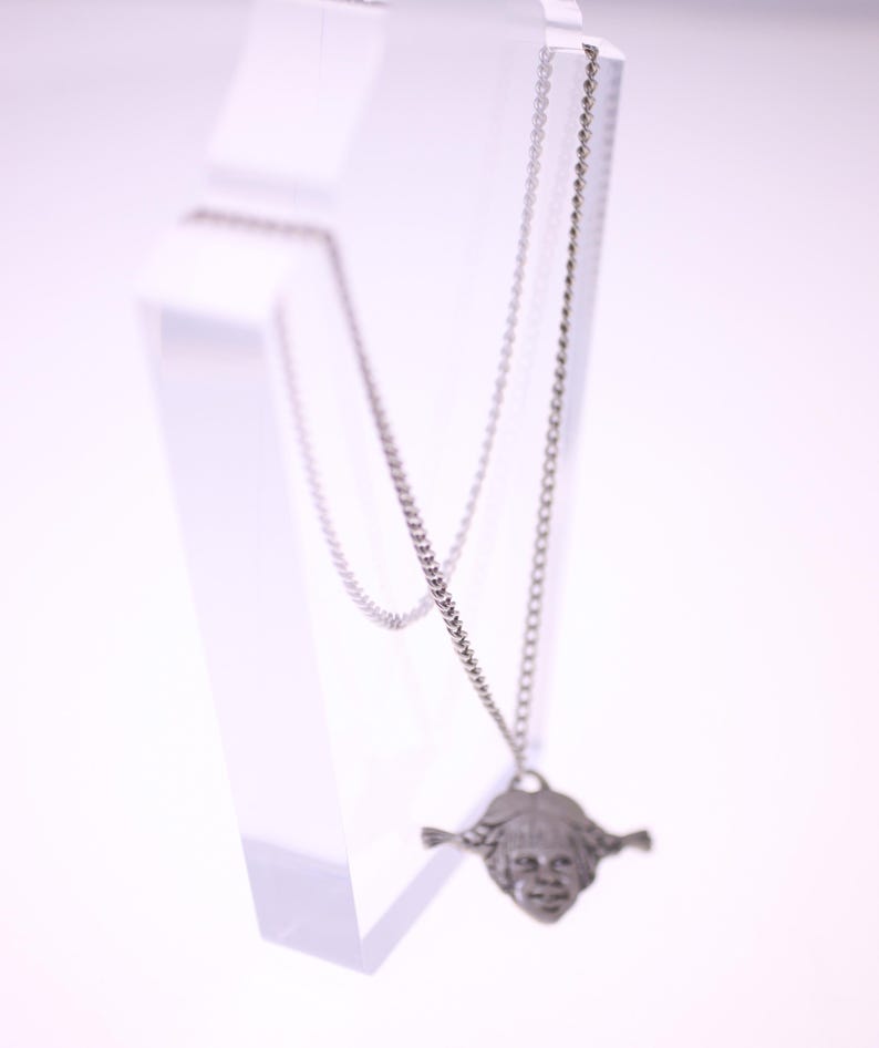 May include: A silver-colored necklace with a pendant shaped like a face with braided hair. The pendant is suspended from a delicate chain. The necklace is displayed on a clear, angled stand against a white background.