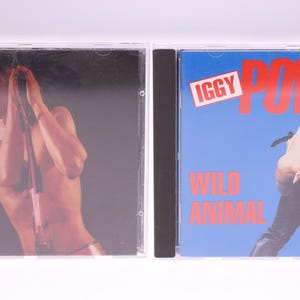 Può includere: Due custodie per CD. La custodia di sinistra presenta una persona che tiene un microfono. La custodia di destra ha il testo "IGGY POP" e "WILD ANIMAL" su sfondo blu. L'immagine a destra mostra una persona con la testa all'indietro.