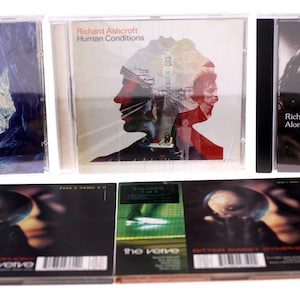 Può includere: Una collezione di CD musicali con copertine di album diverse. I CD includono titoli come "Human Conditions" di Richard Ashcroft e "Alone With Everybody". La fila inferiore presenta CD di "The Verve".
