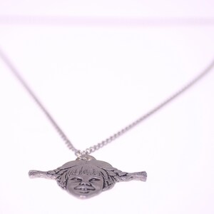 May include: Silver-toned pendant necklace featuring a stylized face with braided pigtails. The pendant is suspended from a delicate chain. The necklace is set against a white background.