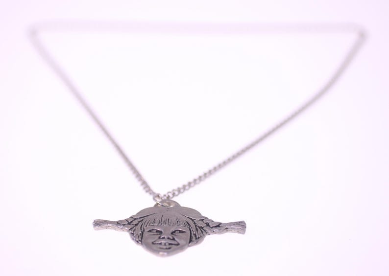May include: Silver-toned pendant necklace featuring a stylized face with braided pigtails. The pendant is suspended from a delicate chain. The necklace is set against a white background.