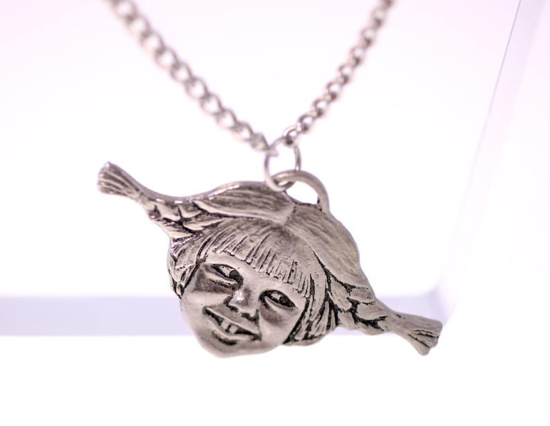 May include: A silver-toned pendant necklace featuring a detailed, smiling face with bangs and side braids. The pendant is suspended from a delicate chain. The necklace is displayed against a bright white background.