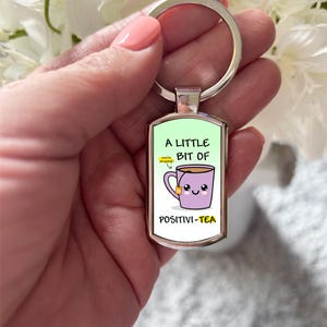 May include: A silver keyring with a green rectangular charm. The charm features a cartoon illustration of a purple teacup with a tea bag and a smiling face. The text on the charm reads "A little bit of Positivi-Tea".
