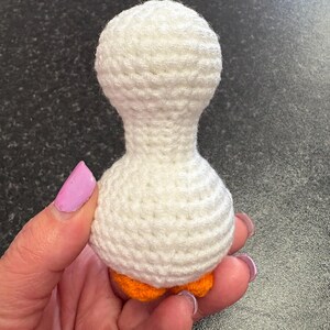 May include: A handmade, white crochet goose with orange feet. The goose is crafted with a textured stitch, featuring a rounded head and body. The small, decorative goose is held in a hand.