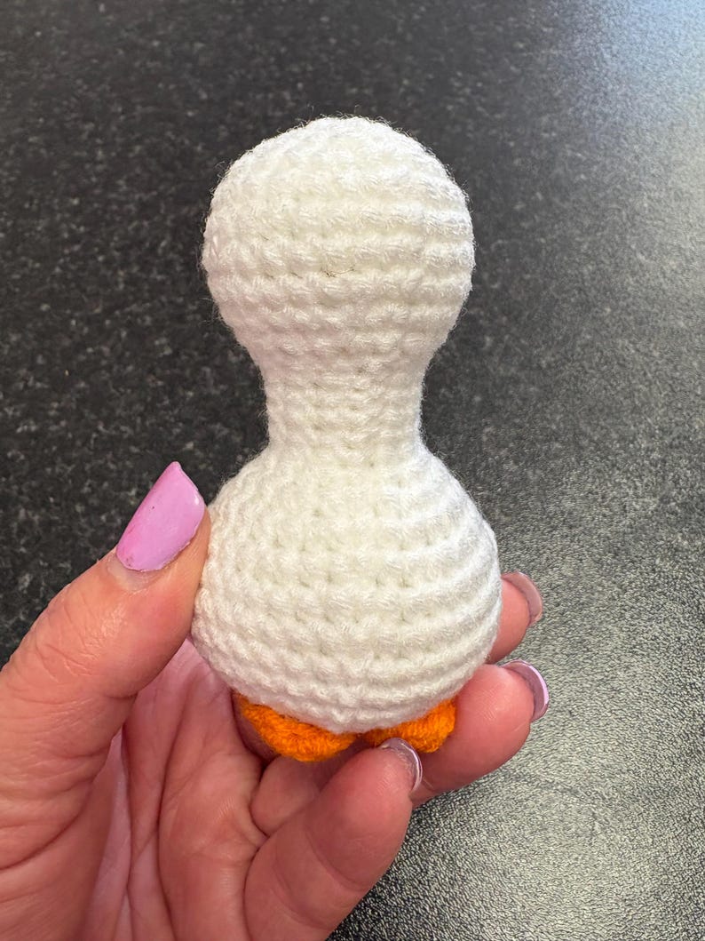 May include: A handmade, white crochet goose with orange feet. The goose is crafted with a textured stitch, featuring a rounded head and body. The small, decorative goose is held in a hand.