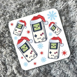May include: White ceramic coaster with a festive design featuring five cartoon game consoles wearing Santa hats. The consoles are surrounded by red stars and blue snowflakes.
