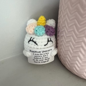 May include: A crocheted white unicorn with a yellow horn and colourful accents. The unicorn has a smiling face with black stitched eyes and a message that reads "Positive Unicorn: I may be a tiny unicorn, but I believe in you. Go do your thing! You're magical!"