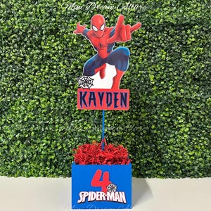 May include: A blue and red centerpiece with a Spider-Man theme. The centerpiece features a red and blue box with the number 4 and the words "Spider-Man". A red and blue cardboard cutout of Spider-Man is attached to a stick and placed in the box. The stick is attached to a red and blue sign with the name "Kayden".