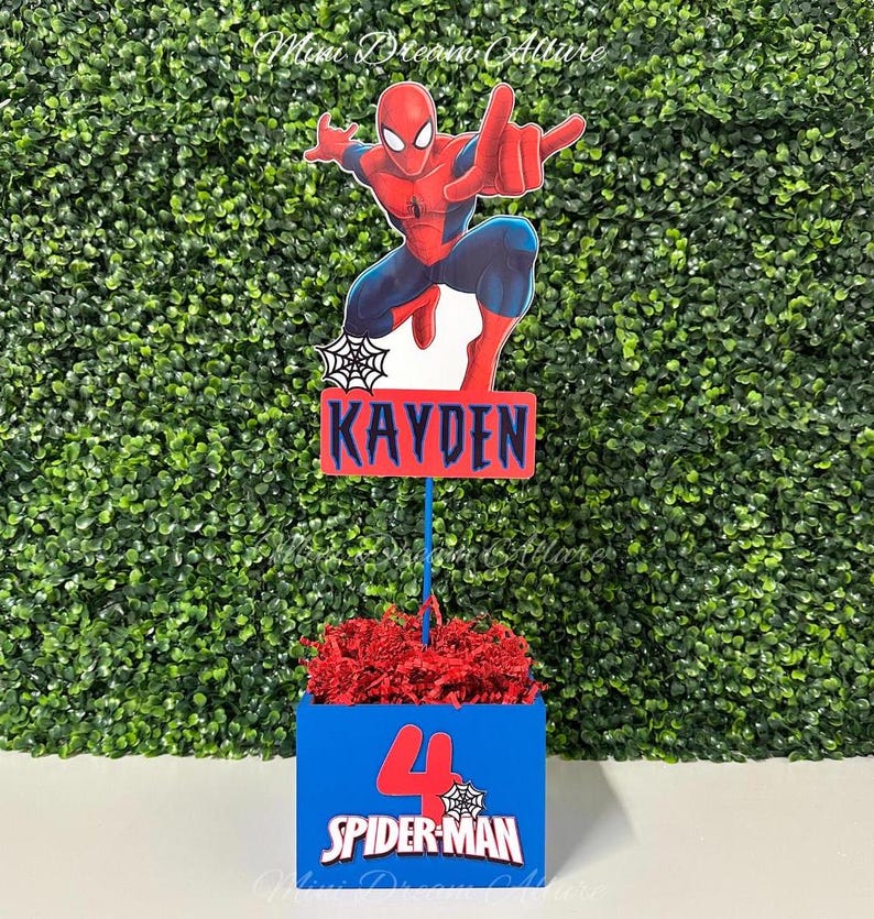 May include: A blue and red centerpiece with a Spider-Man theme. The centerpiece features a red and blue box with the number 4 and the words "Spider-Man". A red and blue cardboard cutout of Spider-Man is attached to a stick and placed in the box. The stick is attached to a red and blue sign with the name "Kayden".