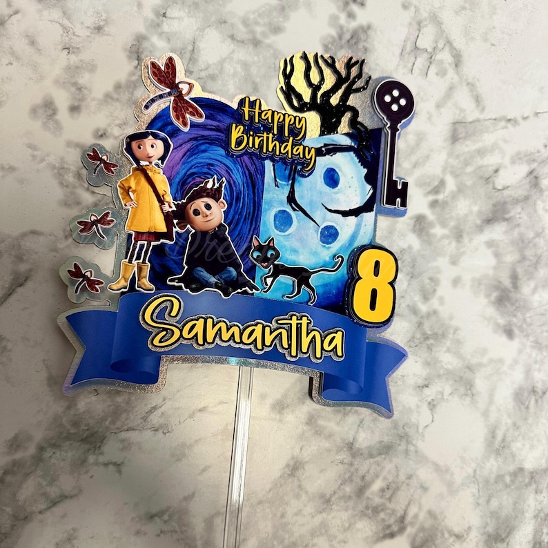 Puede incluir: Un adorno para pastel azul y amarillo con personajes de la pel&iacute;cula "Coraline" con el texto "Feliz cumplea&ntilde;os" y "Samantha" y el n&uacute;mero "8".