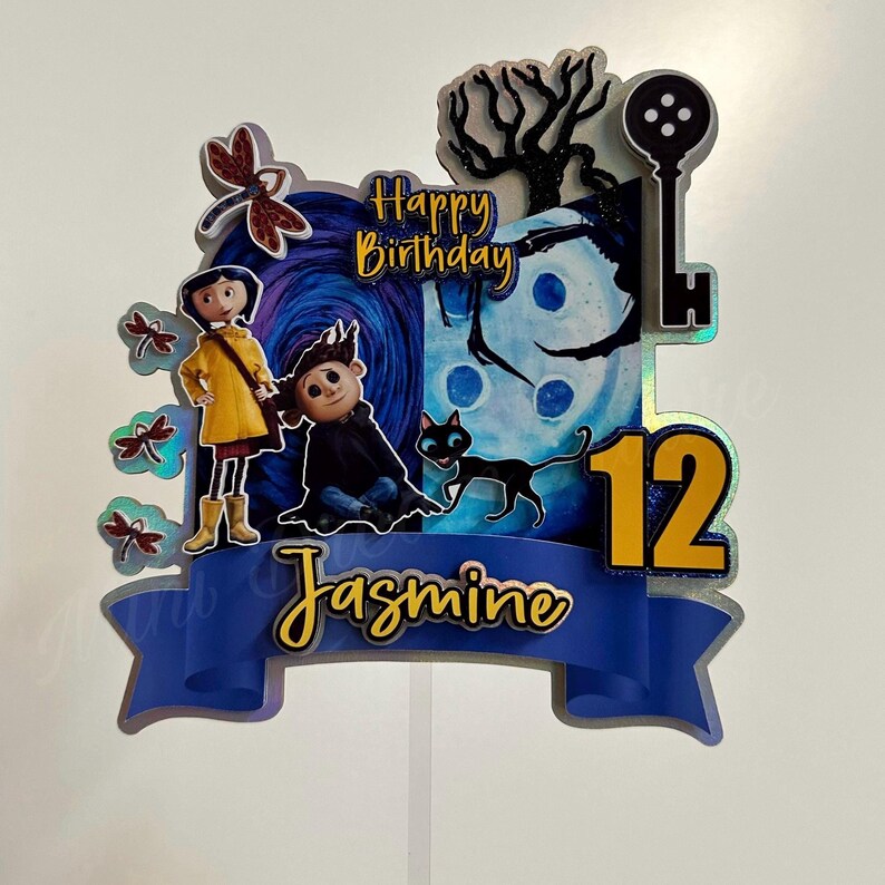 Puede incluir: Un adorno para pastel azul y amarillo con una pancarta azul que dice "Jasmine" y el n&uacute;mero "12". El adorno presenta personajes de la pel&iacute;cula "Coraline" con el texto "Feliz cumplea&ntilde;os".