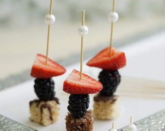 May include: Three skewers with white pearl tops, each with a square of rice crispy treat, a blackberry, a strawberry slice, and a white pearl top.