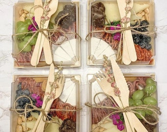Puede incluir: Cuatro pequeñas cajas cuadradas llenas de embutidos y queso. Cada caja está atada con cordel y decorada con flores secas. Las cajas contienen queso, salami, uvas, nueces y galletas.