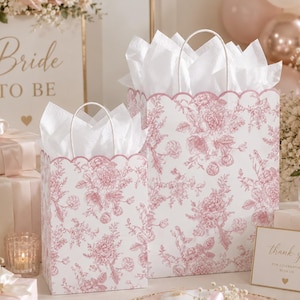 Puede incluir: Dos bolsas de regalo blancas con un patrón floral rosa tipo toile y bordes superiores festoneados, cada una con papel de seda blanco. Las bolsas tienen asas blancas resistentes y se muestran junto a regalos envueltos y un cartel que dice Bride to be.