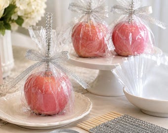 May include: Three red candy apples wrapped in clear cellophane, each topped with a silver rhinestone-encrusted stick and a silver glitter ribbon bow. The apples are displayed on white plates and a cake stand, accompanied by extra sticks and ribbon.