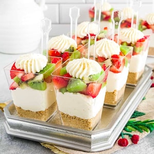 以下が含まれることがあります： 銀色のトレイに、チーズケーキ、ホイップクリーム、キウイ、イチゴの層で満たされた個別のデザートカップが置かれています。カップには透明なプラスチックのスプーンが載せられています。