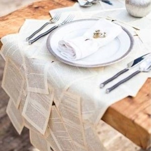 May include: A white table setting with a white tablecloth made of pages from a book. The table is set with white plates, silverware, and napkins. There is a small silver ornament on the table.