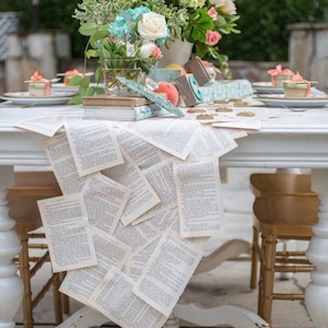 Könnte beinhalten: Ein weißer Holztisch ist mit einem Tischläufer aus überlappenden alten Buchseiten dekoriert. Das Herzstück besteht aus einem Blumenarrangement mit rosa und weißen Rosen, grünen Hortensien, gestapelten Büchern und kleinen Gläsern.