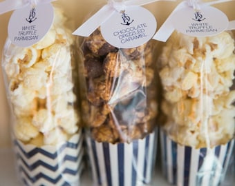 May include: Three clear cellophane bags filled with popcorn and other treats, tied with white ribbon and a circular tag. The tags have a nautical anchor logo and text that reads "White Truffle Parmesan", "Chocolate Drizzled Caramel", and "White Truffle Parmesan". The bags are in striped paper cups.