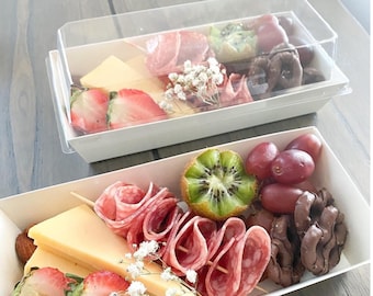 May include: A white cardboard box filled with a charcuterie board featuring salami, cheese, grapes, kiwi, chocolate pretzels, and strawberries.