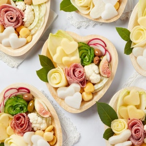 Puede incluir: Una colección de platos en forma de corazón llenos de varios bocadillos, que incluyen queso, salami, coliflor y pepinos. Los platos están dispuestos sobre una superficie blanca con un mantel de encaje.