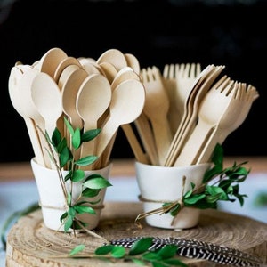 May include: Two white ceramic cups filled with wooden spoons and forks, decorated with green sprigs and twine. The cups are sitting on a wooden slice.