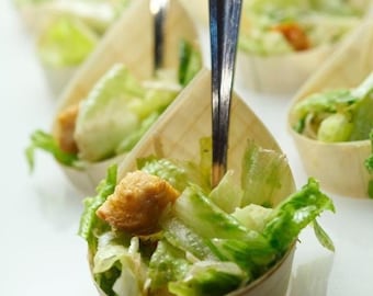Puede incluir: Primer plano de un cuenco de madera lleno de ensalada César con picatostes y lechuga. El cuenco está sobre una superficie blanca con una cuchara de plata que sobresale de la ensalada.