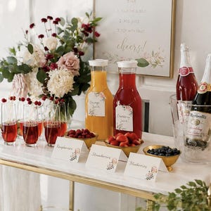 Può includere: Un carrello bar in metallo dorato con piano in marmo bianco, un cartello Mimosa Bar, due caraffe in vetro con succo d'arancia e di mirtillo rosso, tre ciotole dorate con lamponi, fragole e mirtilli, e due bottiglie di sidro frizzante nel ghiaccio.