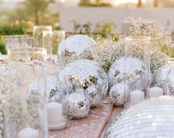 May include: A table setting with silver disco balls, white candles, and white flowers. The table is covered in a light pink tablecloth.