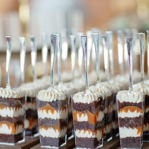 May include: A selection of individual layered desserts in clear cups. The cakes feature layers of chocolate sponge, cream, and caramel, finished with a swirl of cream and a small silver spoon.