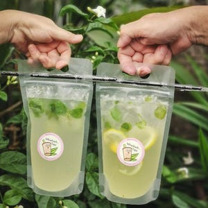 May include: Two clear plastic pouches with a straw hanging out of the top. The pouches contain a light yellow liquid with green mint leaves and lemon slices. The pouches have a pink sticker with the text "Winkwink Tea" and a cartoon teacup with a face.